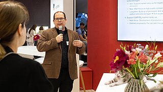 Ein Sprecher des PtX Lab Lausitz hält einen Vortrag auf der ILA Berlin. Er trägt ein braunes Sakko und spricht in ein Handmikrofon. Neben ihm zeigt ein Bildschirm Details zum SAF/PtL-Markthochlauf. Im Vordergrund sitzt ein gähnendes Kätzchen neben einem Blumenstrauß auf einem weißen Tisch.