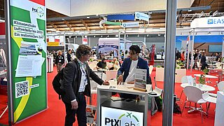 Ein Mitarbeiter des PtX Lab Lausitz im blauen Sakko unterhält sich mit einem Besucher an einem Messestand. Im Vordergrund steht ein großes Banner mit der Aufschrift „Klimaneutraler Luftverkehr bis 2050: Ein realistisches Ziel?“. Der Stand ist mit dem Lab-Logo gebrandet; im Hintergrund ist die Messehalle mit rotem Teppich zu sehen.