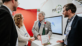 Dr. Harry Lehmann vom PtX Lab Lausitz im Gespräch mit Fachkollegen am Messestand auf der Hannover Messe. Im Hintergrund sind das Lab-Logo, ein Monitor mit Grafiken und ein Hinweisschild auf Publikationen zu sehen.