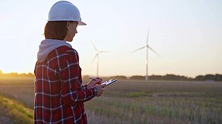 Ingenieurin in karierter Arbeitskleidung macht sich auf einem Feld mit Windkraftanlagen Notizen auf einem Klemmbrett, während die Sonne untergeht, Rückansicht.