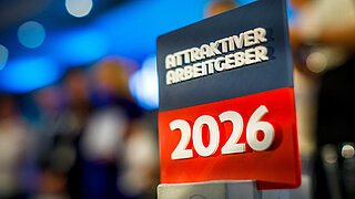 Nahaufnahme der Auszeichnung „Attraktiver Arbeitgeber 2026" vor unscharfem blauen Hintergrund mit Veranstaltungsatmosphäre.