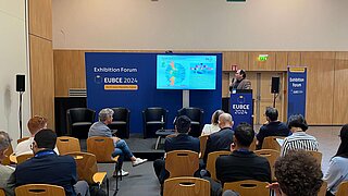 Ein Experte des PtX Lab Lausitz hält einen Vortrag im „Exhibition Forum“ der EUBCE 2024 in Marseille. Er steht an einem blauen Rednerpult vor einem aufmerksamen Publikum; die Leinwand im Hintergrund zeigt eine Folie zu Nachhaltigkeitskriterien und Klimaneutralität.