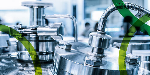 Im Bildfokus das Detail einer chemischen Anlage aus Edelstahl: ein flexibler Segmentschlauch ist durch ein Schraubgewinde mit einem runden Deckel verbunden, der durch vier Sechskantbolzen befestigt ist. Im Hintergrund sind unscharf weitere Edelstahlobjekte sichtbar und durch grafische Nachbearbeitung darübergelegt die grünen gebogenen Linien aus dem Logo von PtXLab Lausitz