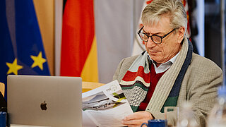Dr. Harry Lehmann vom PtX Lab Lausitz liest konzentriert ein Informationsblatt während einer Veranstaltung in Brüssel. Er sitzt an einem Tisch mit einem Laptop und einer blauen Tasse; im Hintergrund sind die Flaggen der Europäischen Union und Deutschlands zu sehen.