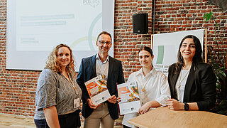 Vier Teammitglieder des PtX Lab Lausitz präsentieren neue Publikationen bei einer Veranstaltung in Brüssel. Sie stehen vor einer Backsteinwand; im Hintergrund zeigt eine Leinwand den Titel „The raw materials behind synthetic kerosene – Securing critical resources for the EU“. Zwei Personen halten gedruckte Studien zum Rohstoffbedarf für den klimaneutralen Luftverkehr in den Händen.