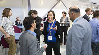 Eine Frau steht in einem Kreis mit Männern in einer Halle und spricht zu Ihnen.