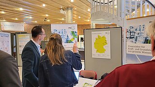 Fachbesucher am Messestand des PtX Lab Lausitz betrachten eine Karte zu PtL-Anlagen in Deutschland. Im Hintergrund sind Informationsbanner und die Architektur der Veranstaltungshalle mit einer markanten Wendeltreppe erkennbar.