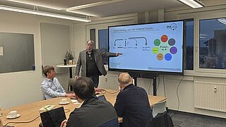Dr. Harry Lehmann hält einen Vortrag vor einer Delegation im PtX Lab Lausitz. Er zeigt auf einen großen Monitor, der eine Grafik zum Thema „Future energy and raw material supply“ und „Power-to-X“ darstellt. Die Zuhörer sitzen an einem langen Holztisch.