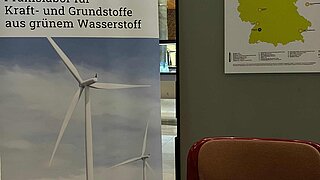 Informationsmaterial des PtX Lab Lausitz auf einem Messestand: Auf einem weißen Tisch liegen Factsheets zum Thema Rohstoffmix. Im Hintergrund sind ein Roll-up-Banner und eine Karte der PtL-Anlagen in Deutschland zu sehen.