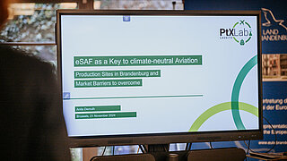 Ein Monitor zeigt die Titelfolie einer Präsentation von Anita Demuth in Brüssel am 21. November 2024. Der Titel lautet „eSAF as a Key to climate-neutral Aviation – Production Sites in Brandenburg and Market Barriers to overcome“. Oben rechts ist das Logo des PtX Lab Lausitz zu sehen.