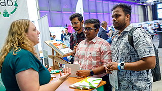 Eine Mitarbeiterin des PtX Lab Lausitz berät drei interessierte junge Männer an einem Messestand während der deCarbonDays. Auf dem Counter liegen Informationsbroschüren aus.