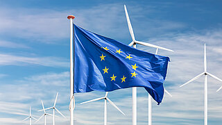 Wolkiger Himmel mit weißen Windrädern. Davor die Flagge der Europäischen Union.