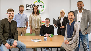 There are seven people in the room. In the centre is a light-coloured table. In the background is the PtX Lab Lausitz logo.