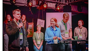 Ein junger Mann namens Lukas spricht bei einem Barcamp in ein Mikrofon. Er steht auf einer Bühne neben weiteren Teilnehmenden (Marika, Simon und Vincent), die alle Lanyards der ZUG tragen. Im Hintergrund sind rote Bühnenstrahler zu sehen.