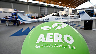 Green balloon with ‘AERO’ logo in an exhibition hall. A light aircraft is in the background.