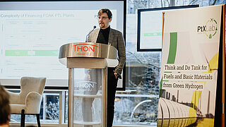 Ein junger Experte mit Brille und Rollkragenpullover hält einen Vortrag an einem Pult im THON Hotel EU. Die Präsentationsfolie im Hintergrund thematisiert die Finanzierung von PTL-Anlagen. Rechts steht ein Banner des PtX Lab Lausitz.