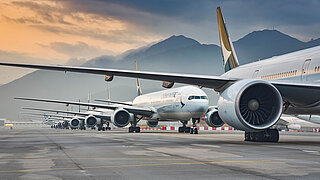 Photo shows several aircrafts parked in a row on a tarmac against a mountain backdrop.