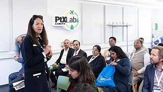 Eine Frau hält ein Mikrofon und spricht zu einer Gruppe von sitzenden Personen, die in einem Raum versammelt sind. An der Wand ist das Logo des PtX Lab Lausitz.