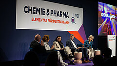 Mehrere Personen stehen dichtgedrängt vor einer Bühne. Auf der Bühne stehen drei Personen. Die Person in der Mitte hält ein Mikrofon uns spricht zum Publikum.