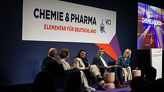 Mehrere Personen stehen dichtgedrängt vor einer Bühne. Auf der Bühne stehen drei Personen. Die Person in der Mitte hält ein Mikrofon uns spricht zum Publikum.