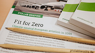 Detailaufnahme eines PtX Lab Briefings mit dem Titel „Fit for Zero – Climate-neutral European aviation in 2050“. Im Vordergrund liegen zwei Bleistifte quer über dem Dokument; rechts sind weitere gestapelte Broschüren zu sehen. Auf dem Briefing ist das Foto eines Flugzeugs abgebildet.