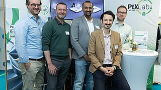 Gruppenfoto von fünf männlichen Personen an einem Messestand.