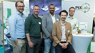 Gruppenfoto von fünf männlichen Personen an einem Messestand.