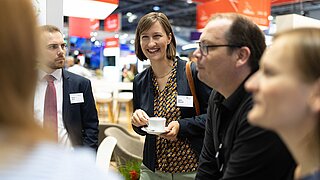 Anita Demuth und Martin Hock vom PtX Lab Lausitz im fachlichen Austausch auf der ILA Berlin 2024. Frau Demuth lächelt und hält eine Kaffeetasse; im Hintergrund ist das geschäftige Treiben der Messe zu sehen.
