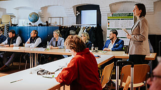 Eine Referentin im beigen Blazer steht und spricht vor Teilnehmenden, die an langen Tischen sitzen. Im Hintergrund ist ein wissenschaftliches Poster zur Nachhaltigkeitsanalyse von Kraftstoffen aus grünem Wasserstoff zu sehen.