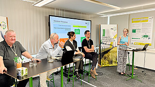 Five people in the foreground at bar tables. Behind them is a screen showing a presentation, along with posters and display stands.