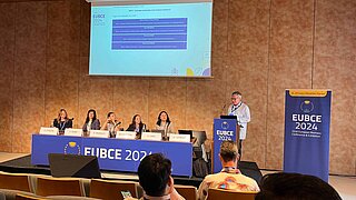 Dr. Harry Lehmann vom PtX Lab Lausitz moderiert eine Sitzung bei der „EUBCE 2024“ (European Biomass Conference & Exhibition) in Marseille. Er steht an einem blauen Rednerpult neben einem Podium mit fünf Expertinnen. Die Leinwand zeigt das Thema „Hydrogen production from biomass feedstock“.