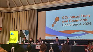 Podiumsdiskussion bei der „CO2-based Fuels and Chemicals Conference 2024“. Fünf Experten sitzen auf einer Bühne vor einer großen Projektionsfläche mit dem Veranstaltungstitel. Links stehen Banner von Nova und Yncoris.