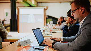 Christopher Kupke from PtX Lab Lausitz works on his laptop during the Maritime Round Table in Hamburg. A nameplate and a recycled paper notepad are on the wooden table in front of him.