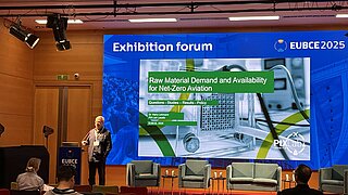 Dr. Harry Lehmann vom PtX Lab Lausitz hält einen Vortrag auf der EUBCE 2025. Die große Leinwand zeigt den Titel „Raw Material Demand and Availability for Net-Zero Aviation“. Im Vordergrund sitzen Zuschauer vor der Bühne mit leeren Sesseln.