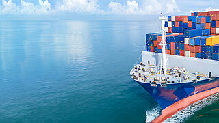 Luftaufnahme eines großen Frachtschiffs, das vollständig mit bunten Schiffscontainern beladen ist und durch ruhiges Meerwasser fährt.