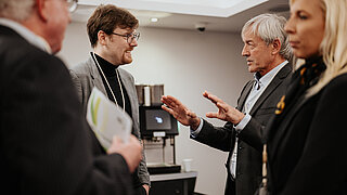 Vier Personen im Gespräch bei einer Fachveranstaltung in Brüssel. Ein älterer Mann im grauen Anzug gestikuliert lebhaft, während er mit einem jüngeren Kollegen mit Brille spricht. Im Hintergrund steht eine Kaffeemaschine.