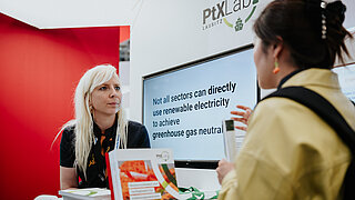 Eine blonde Expertin des PtX Lab Lausitz im Gespräch mit einer Besucherin am Messestand auf der Hannover Messe 2025. Hinter ihr zeigt ein Monitor den englischen Satz: „Not all sectors can directly use renewable electricity to achieve greenhouse gas neutrality.“ Auf dem Tresen liegen Informationsbroschüren.