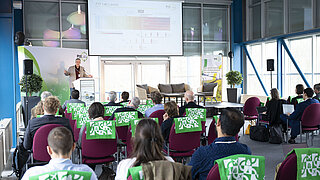 Redner steht vor einem Rednerpult und spricht zum Publikum in einem Konferenzraum.