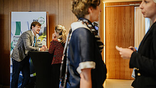 Ein Mann unterhält sich mit einer Frau an einem Konferenzstand und weiter rechts unterhalten sich zwei Frauen
