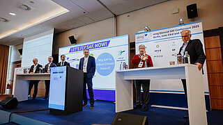 Sechs Männer stehen auf einer Bühne und einer von ihnen spricht