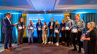 Gruppenaufnahme mehrerer Preisträger*innen bei der Verleihung des Titels „Attraktiver Arbeitgeber 2026" in Cottbus. Die Ausgezeichneten stehen auf einer Bühne vor einer blauen Leinwand und halten ihre Plaketten in die Kamera.