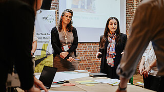 Zwei Expertinnen des PtX Lab Lausitz stehen an einem großen Arbeitstisch während eines Workshops in Brüssel am 13. Mai 2025. Eine Frau im dunklen Blazer und gemustertem Schal gestikuliert erklärend, während ihre Kollegin im braunen Oberteil aufmerksam zuhört. Im Hintergrund sind eine Backsteinwand, eine Präsentationsleinwand und ein Roll-up-Banner des PtX Lab zu erkennen.