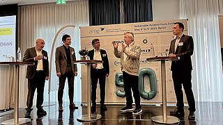 Fünf Personen stehen auf einer Bühne. Eine Person spricht hält ein Mikrofon und spricht. Im Hintergrund eine Presswand mit Logos.