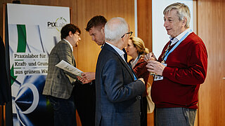 Ein älterer Mann im roten Pullover unterhält sich bei einer Fachveranstaltung mit Kollegen vor einem Roll-up Banner des PtX Lab Lausitz. Das Banner zeigt Text über Kraftstoffe aus grünem Wasserstoff.