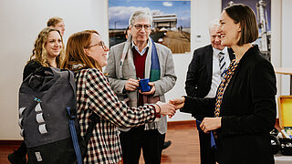 Anita Demuth vom PtX Lab Lausitz begrüßt eine Teilnehmerin herzlich mit einem Händedruck bei einer Veranstaltung in Brüssel. Im Hintergrund steht Dr. Harry Lehmann mit einer blauen Tasse; an der Wand hängen großformatige Bilder von Industrie- und Hafenanlagen.