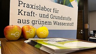 Einige Äpfel mit dem Logo des PtX lab Lausitz liegen auf einem Tisch vor einem Plakat des Labs.