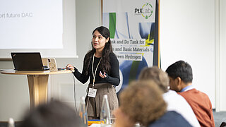 Eine junge Frau mit langen dunklen Haaren steht während eines Vortrags neben einem Holz-Stehpult mit Laptop vor einem Banner des PtX Lab Lausitz und einer Projektionswand mit der Aufschrift „future DAC“, während im unscharfen Vordergrund Zuhörende sitzen.