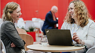 Zwei Frauen im angeregten Gespräch an einem kleinen runden Tisch auf der Hannover Messe 2025. Beide lachen herzlich; auf dem Tisch steht ein geöffneter Laptop. Der Hintergrund des Loungebereichs ist in kräftigem Rot gehalten.