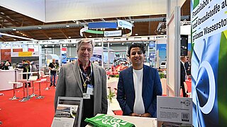 Dr. Harry Lehmann und ein Kollege des PtX Lab Lausitz stehen lächelnd an ihrem Messestand hinter einem weißen Tresen. Auf dem Tisch liegen grüne Stoffbeutel mit Lab-Logo sowie Informationsflyer. Der Hintergrund zeigt das geschäftige Treiben einer Messehalle mit rotem Teppich.