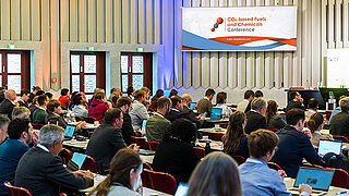 Viele Personen sitzen in einem Saal und verfolgen einen Vortrag.